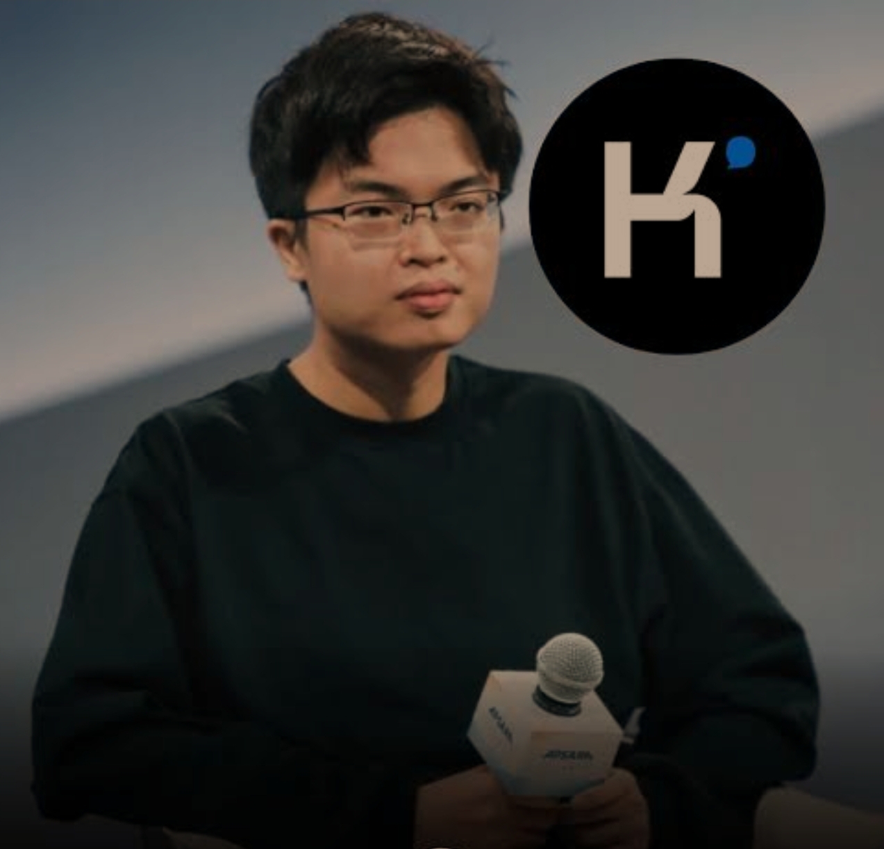 A person holding a microphone is seated with the 'K' logo visible in the background.