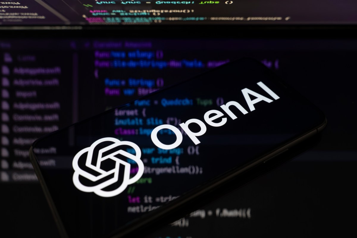 The OpenAI logo is displayed on a smartphone screen placed on a reflective surface onto which lines of computer code.